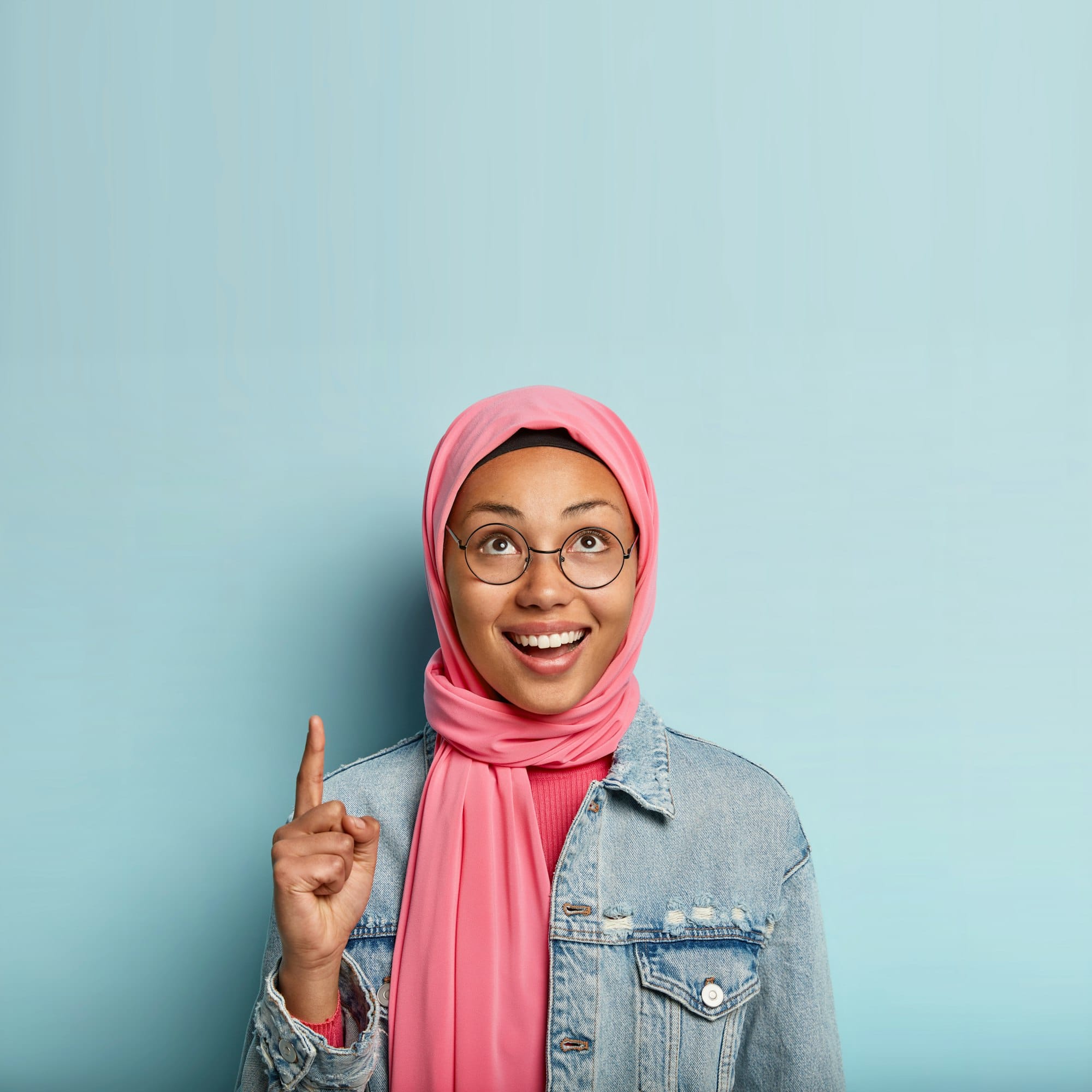 Vertical shot of happy dark skinned female Muslim woman poses over blue background, points above, we with Google Review Management Service Malaysia